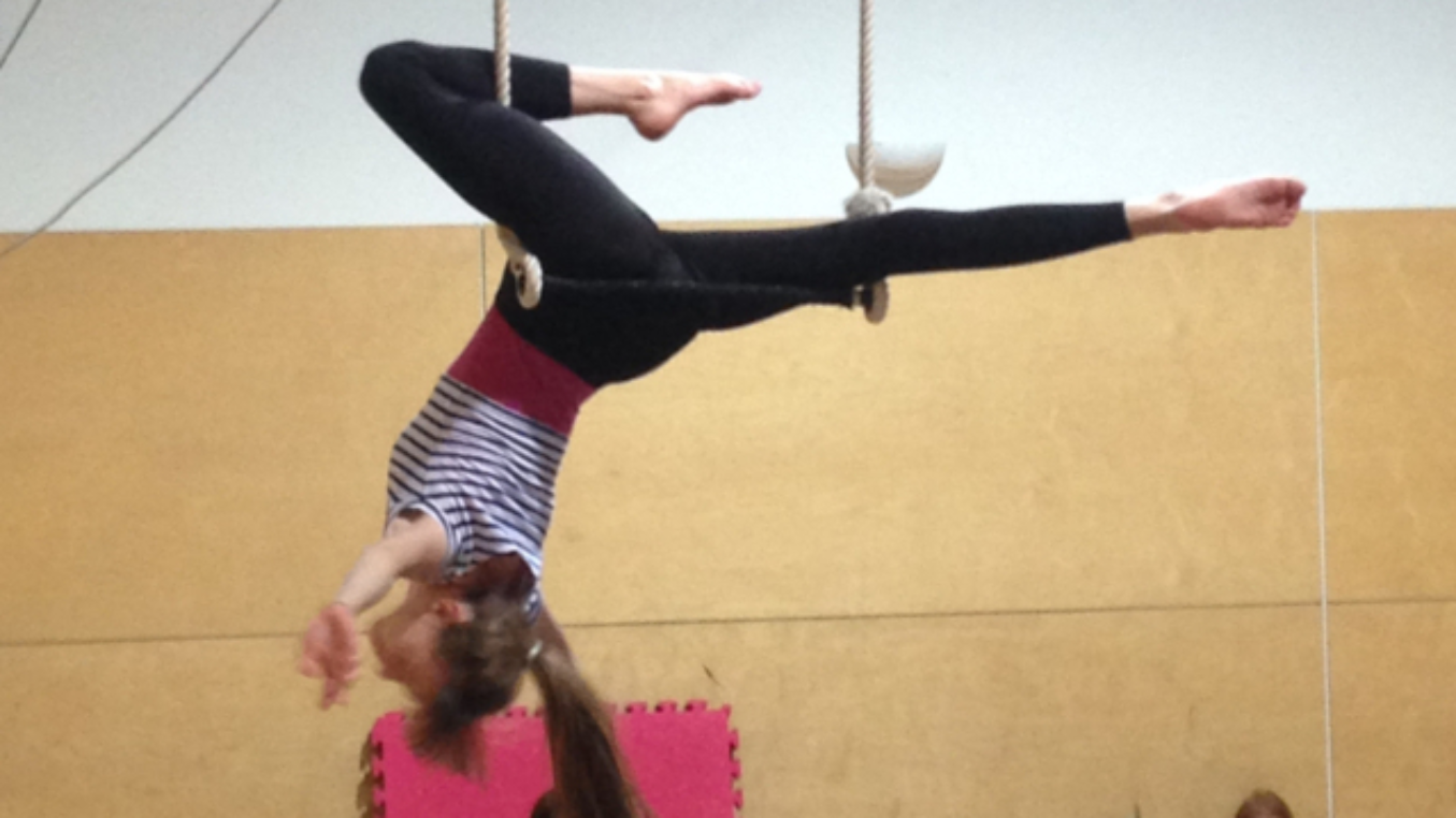 A teenage girl is balancing upside down on a trapeze with one leg bent up and the other straight out behind her. We have a side on view of her and there are children looking up at her.