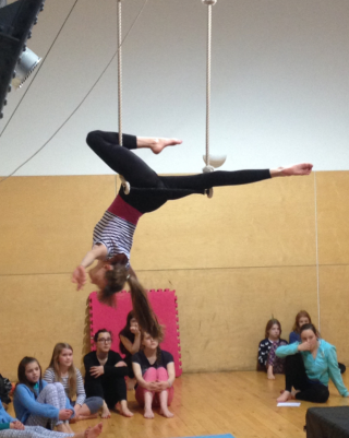 A teenage girl is balancing upside down on a trapeze with one leg bent up and the other straight out behind her. We have a side on view of her and there are children looking up at her.