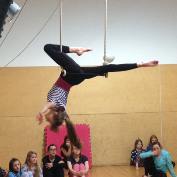 A teenage girl is balancing upside down on a trapeze with one leg bent up and the other straight out behind her. We have a side on view of her and there are children looking up at her.