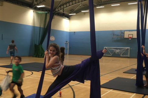 A smiling young girl with hair in a ponytail is suspended in splits in the air on purple silks. She is in a sports hall with a crash mat under her.