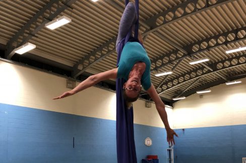 A white woman in sports clothes is hanging upside down on a purple aerial silks with her arms wide. The silks are purple and they are in a sports hall.