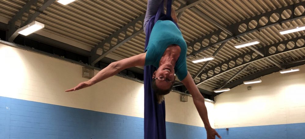 A white woman in sports clothes is hanging upside down on a purple aerial silks with her arms wide. The silks are purple and they are in a sports hall.