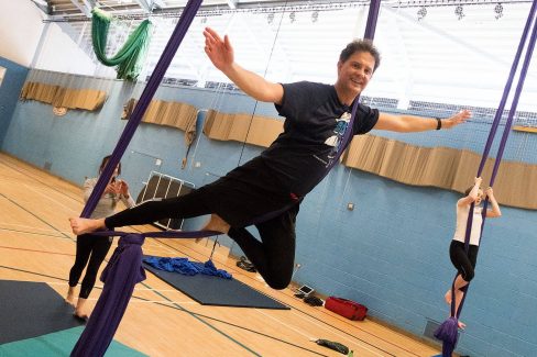 A sports hall with aerial silks hanging down. A man is supsended in one and his hands are free, balancing in the air. There are other people on the other silks in the background