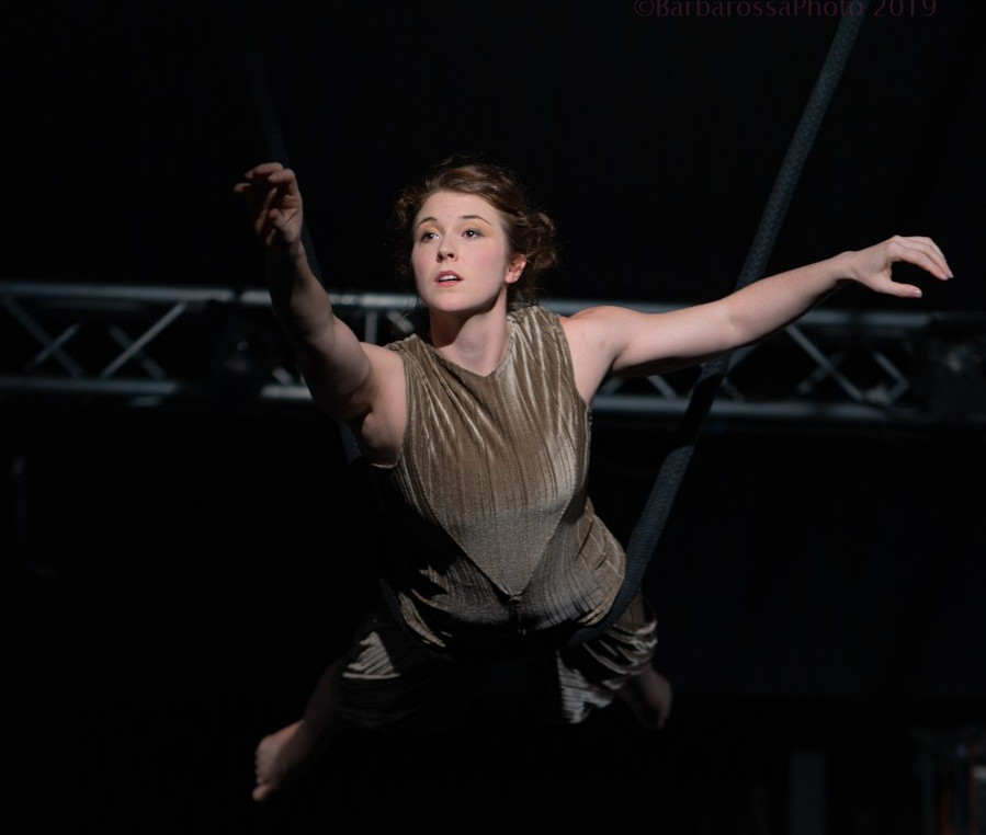 An aerial rope performer mid-perofrmance, reaching her arm forward towards an audience