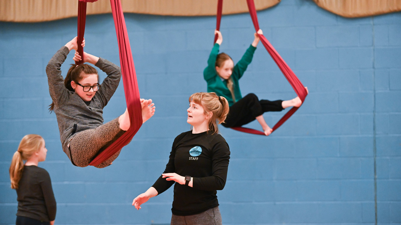 Two junior students making a horizontal shapes in cocoon watched by a teacher.