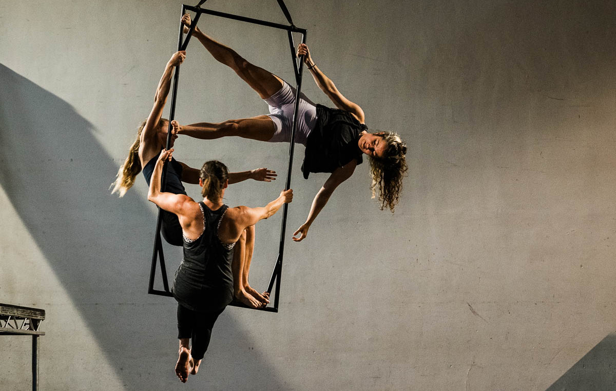 3 aerialists are hanging in and from a rectangular black metal frame representing a window frame. They are on a stage with shadows falling around them.