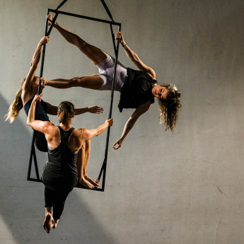 3 aerialists are hanging in and from a rectangular black metal frame representing a window frame. They are on a stage with shadows falling around them.