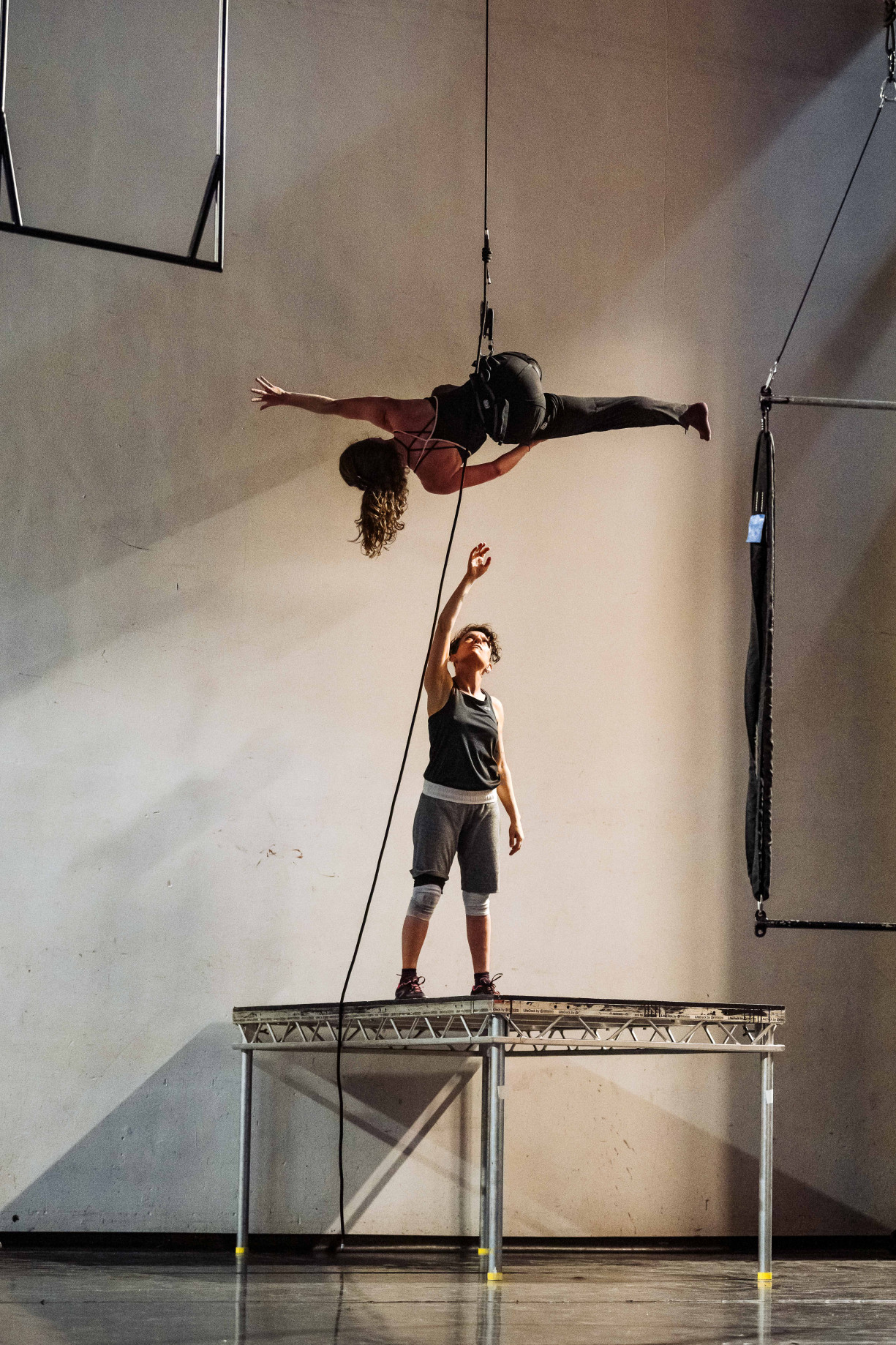 A woman is standing on a raised platform on a stage reaching up to a 2nd performer who is in a harness. This 2nd performer is reaching long, vertical to the floor