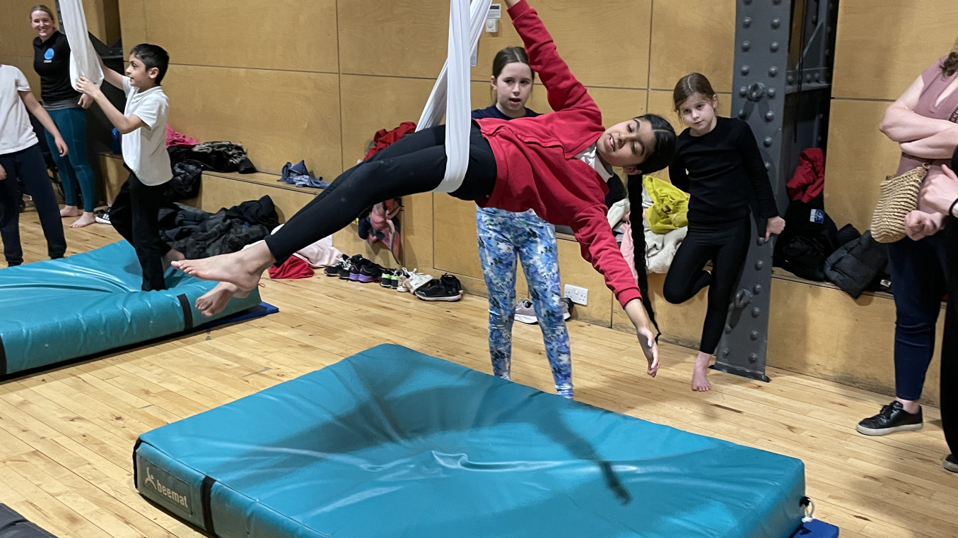 A young girl in a red jumper lies in an aerial sling with one arm up holding the silk and the other arm stretched out to her side.