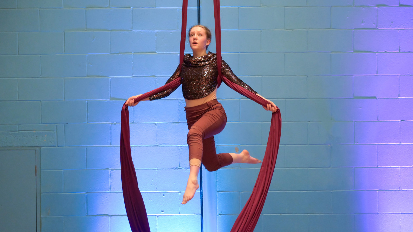 White female youth aerialist is suspended using only her arms out to the side on red silks, her legs are free beneath her. She wears a gold costume and her face is looking out beyond the camera.
