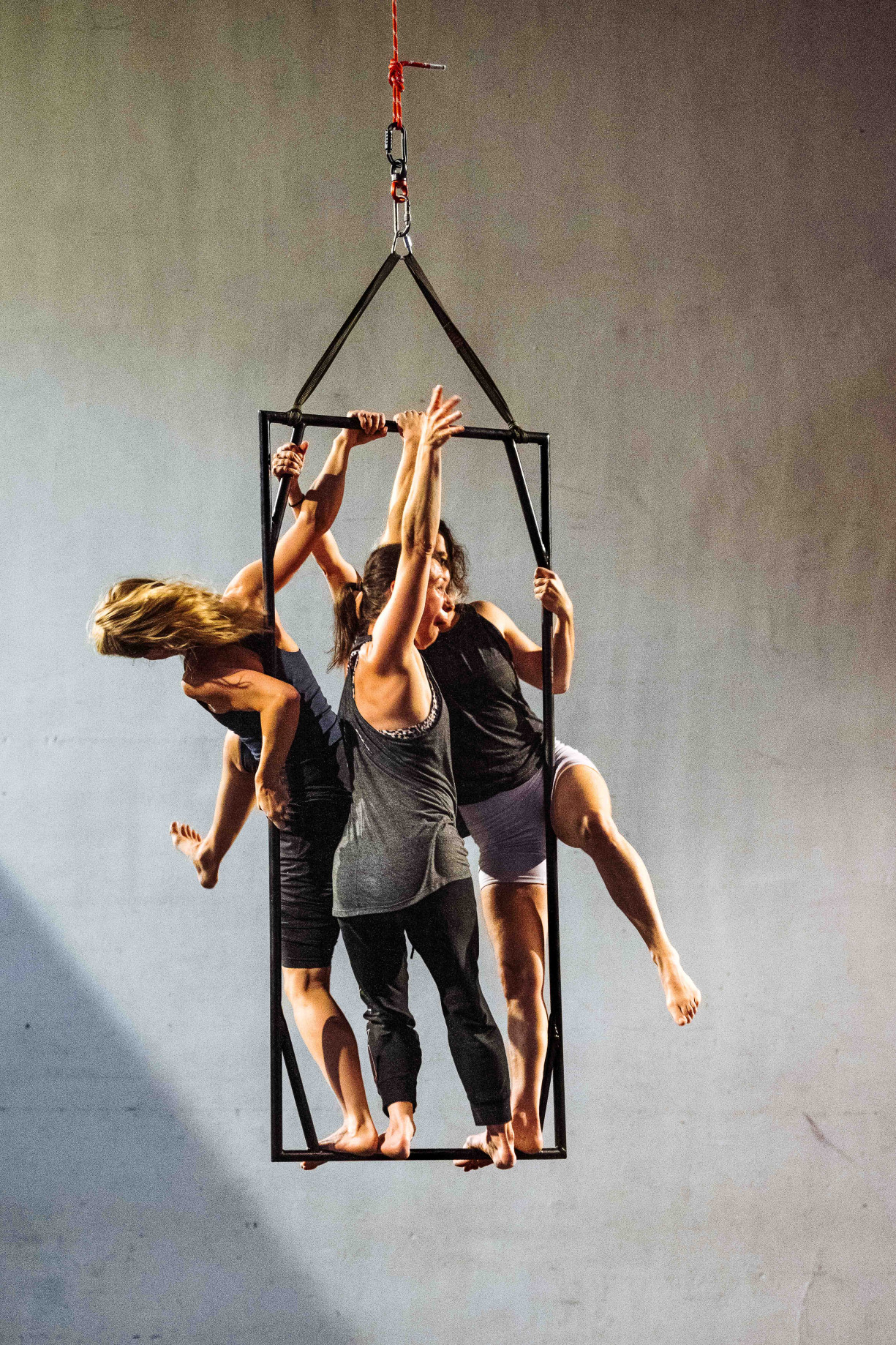3 performers are standing on a black metal frame that is suspended in the air. They are celebrating whilst trying to climb around and over each other