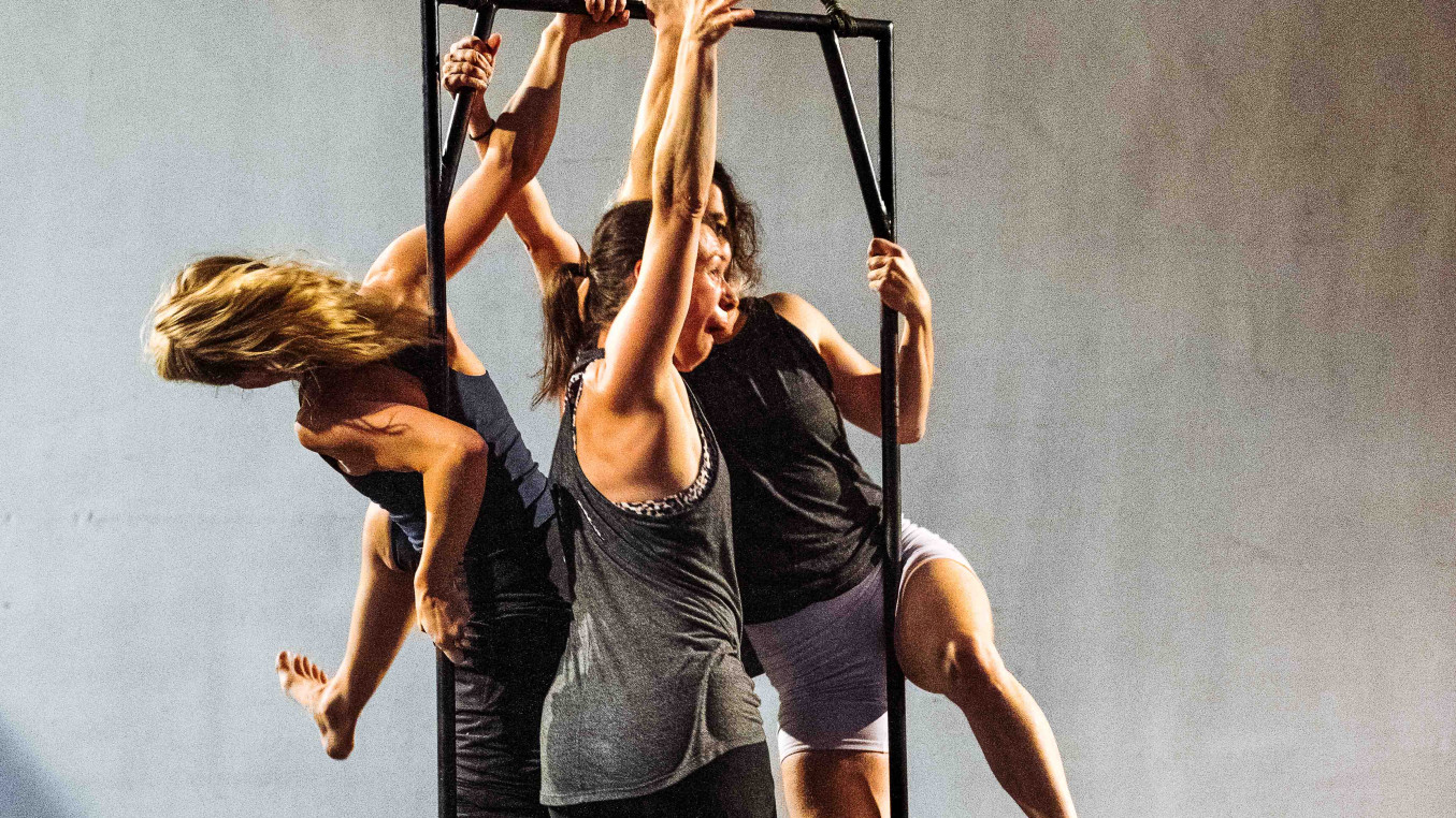 3 performers are standing on a black metal frame that is suspended in the air. They are celebrating whilst trying to climb around and over each other