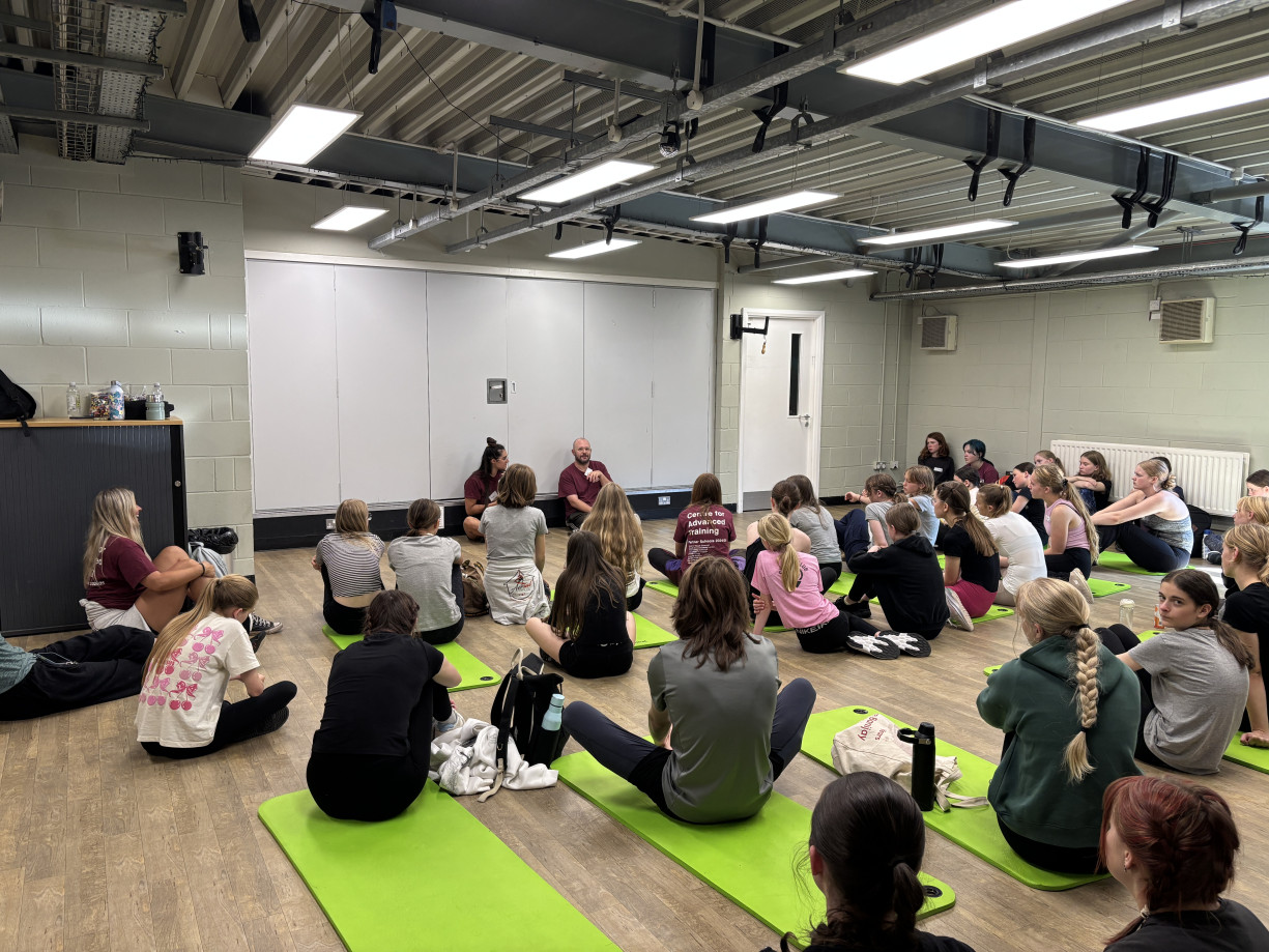 A group of young people are sitting in a room on the floor, listening to 2 people talk