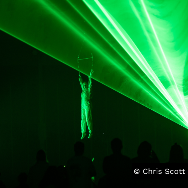 Aerialist is suspended in midair, hanging from a trapeze, while green lasers illuminate the foreground