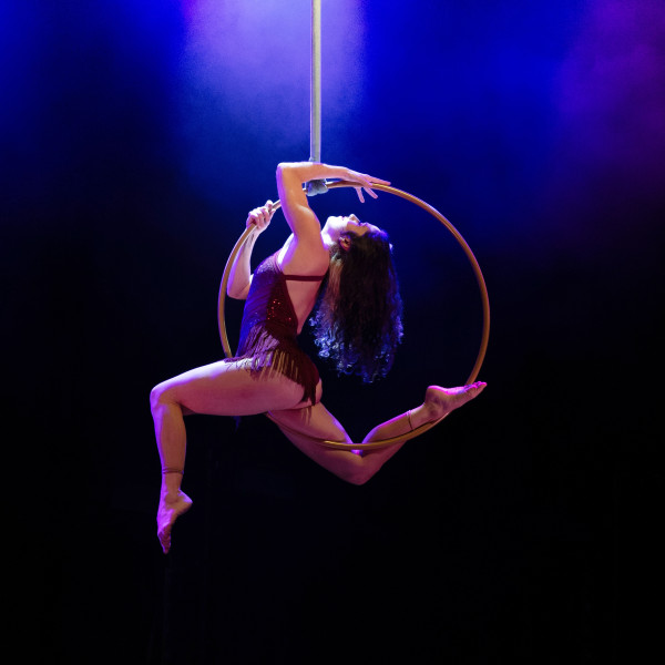 Chrissie Ardill an aerial hoop performer is suspended in an aerial hoop with stage lights shining down on her, she has bent knees in a stag leap shape and she's arching her back to look up. She wears a leotard costume and has long curly hair.