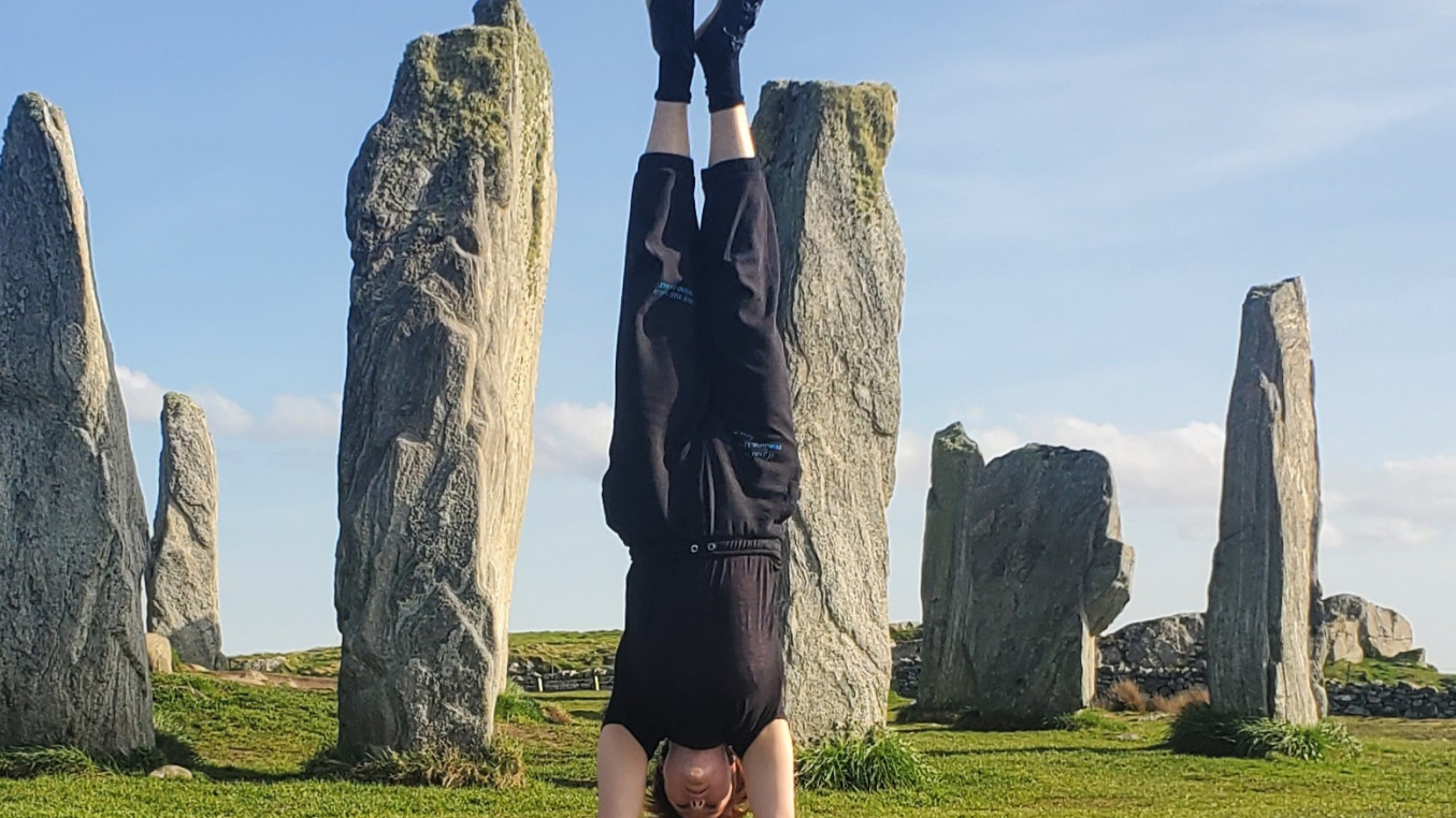 Kit is holding a straight handstand in front of a collection of tall standing stones. They are wearing black trousers and a top. They have short red hair.