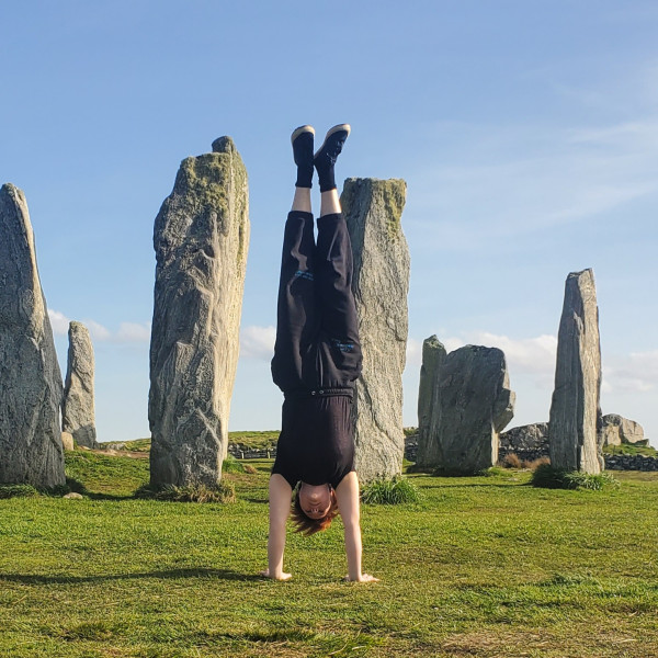 Kit is holding a straight handstand in front of a collection of tall standing stones. They are wearing black trousers and a top. They have short red hair.