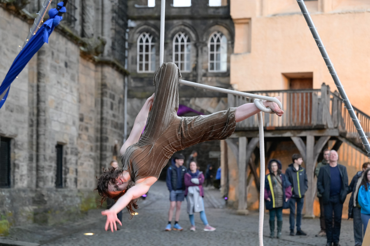A woman is upside down on a rope with one leg hooked on to it and the other wrapped in a knot. She is reaching with one hand towards the camera. She is outside in the grounds of Stirling castle