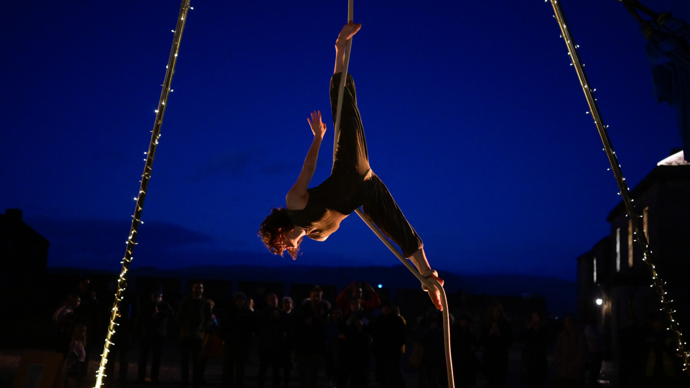 A woman is upside down on a rope in an acrobatic pose. It is dark and she is lit by fairy lights that hang around the poles of an aerial rig