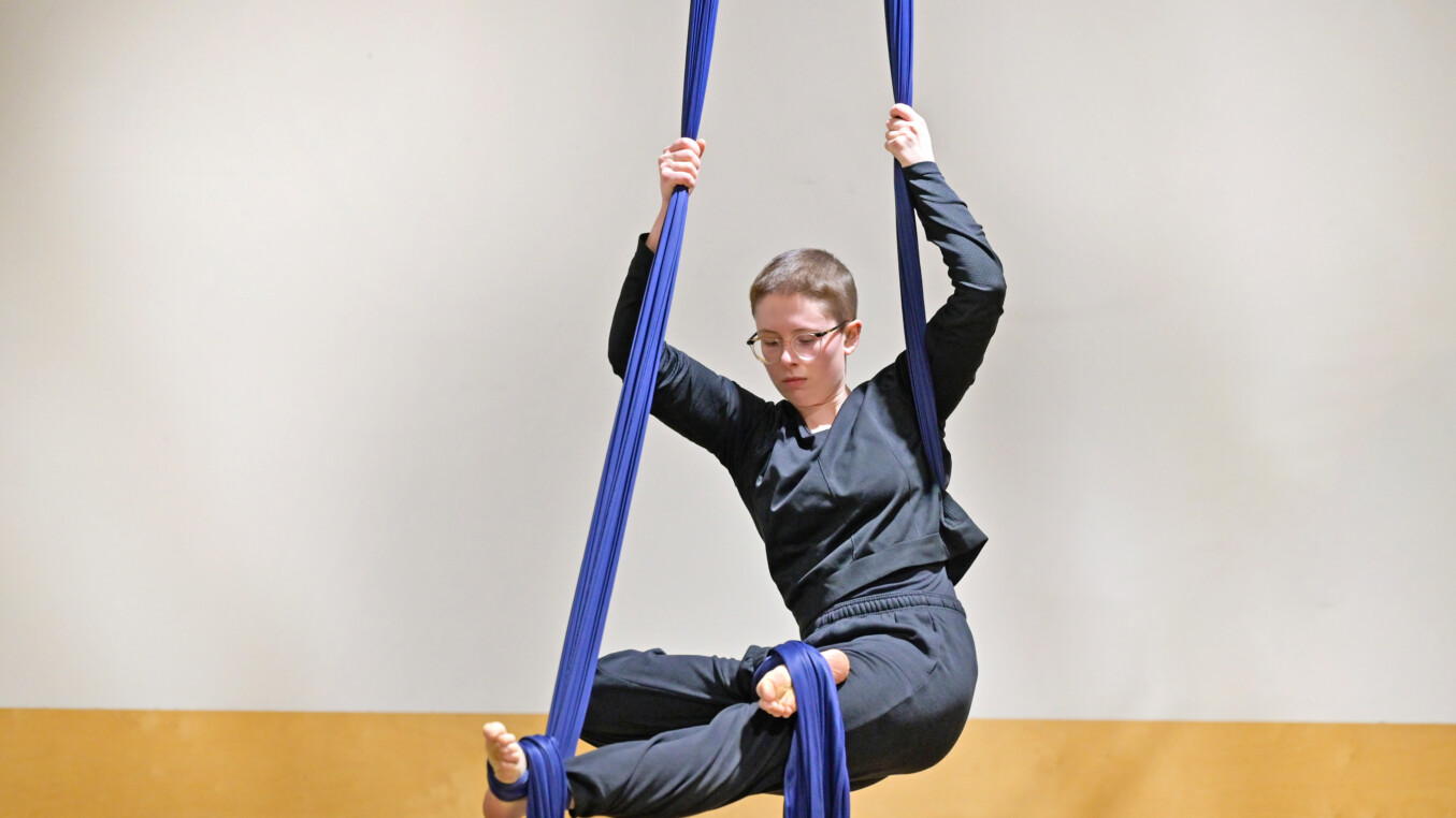 An aerialist with an aerial silk footlocked around each foot sinks into a seated position against one side of the silk with it tucked into her underarm and wrapped around her waist.