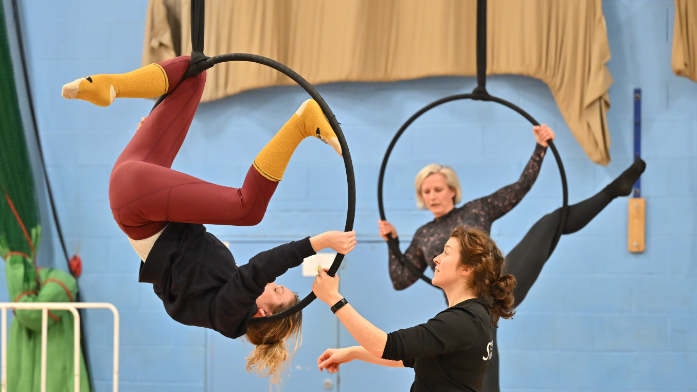 three women are in the image, 2 are in the air in aerial hoops, one of whom is upsdie down. The third woman is on the floor instructing the one upside down. They are wearing training clothes and are in a blue painted sports hall