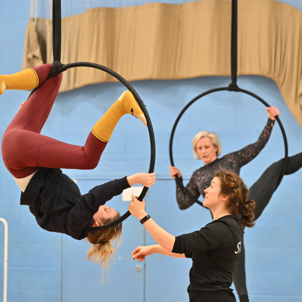 three women are in the image, 2 are in the air in aerial hoops, one of whom is upsdie down. The third woman is on the floor instructing the one upside down. They are wearing training clothes and are in a blue painted sports hall