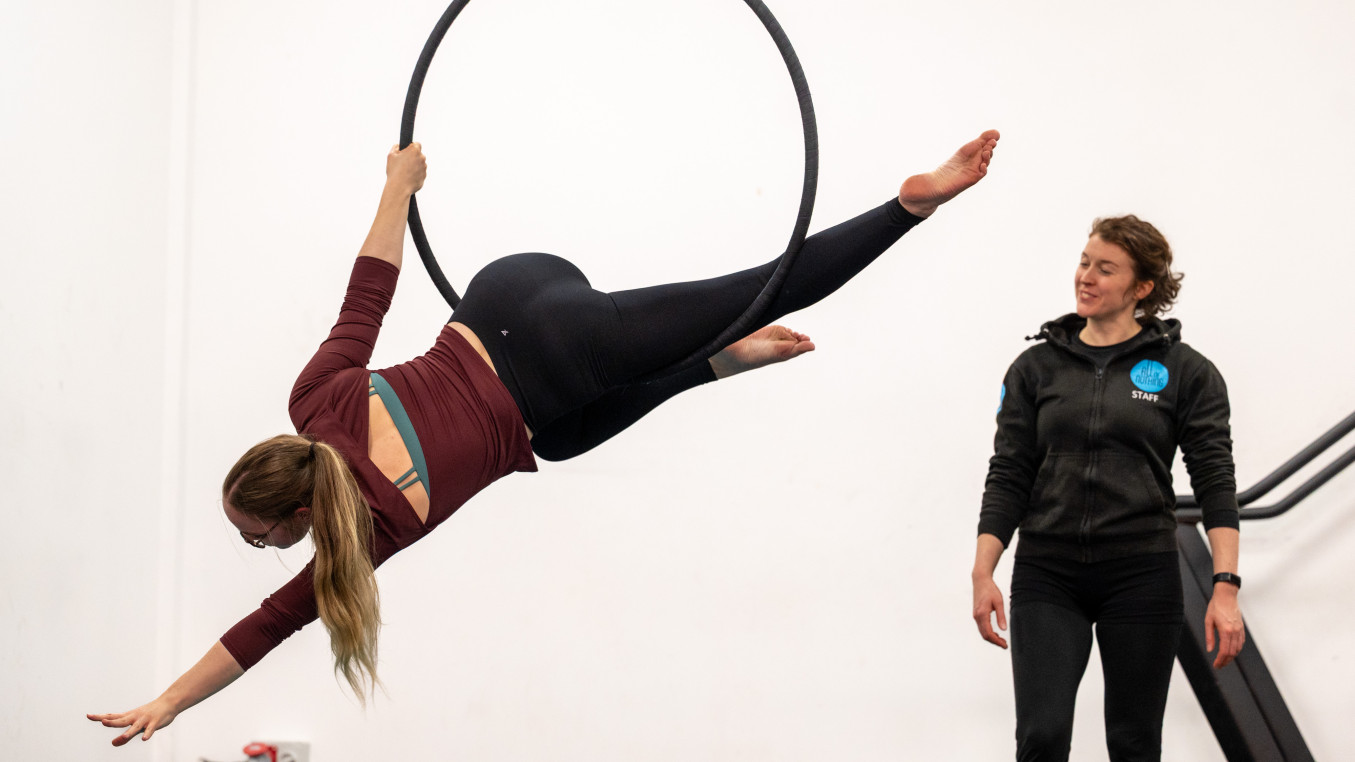 Two women are in the picture, the one on the right is suspended in an aerial hoop. She is on her left side and reaching diagonally down towards the floor. Her back is facing the camera. The second woman is facing her standing to the right side of the image and looking on, teaching her.