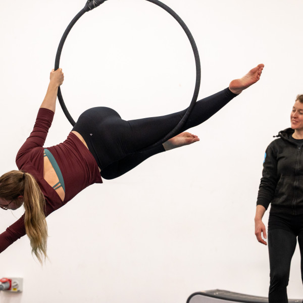 Two women are in the picture, the one on the right is suspended in an aerial hoop. She is on her left side and reaching diagonally down towards the floor. Her back is facing the camera. The second woman is facing her standing to the right side of the image and looking on, teaching her.