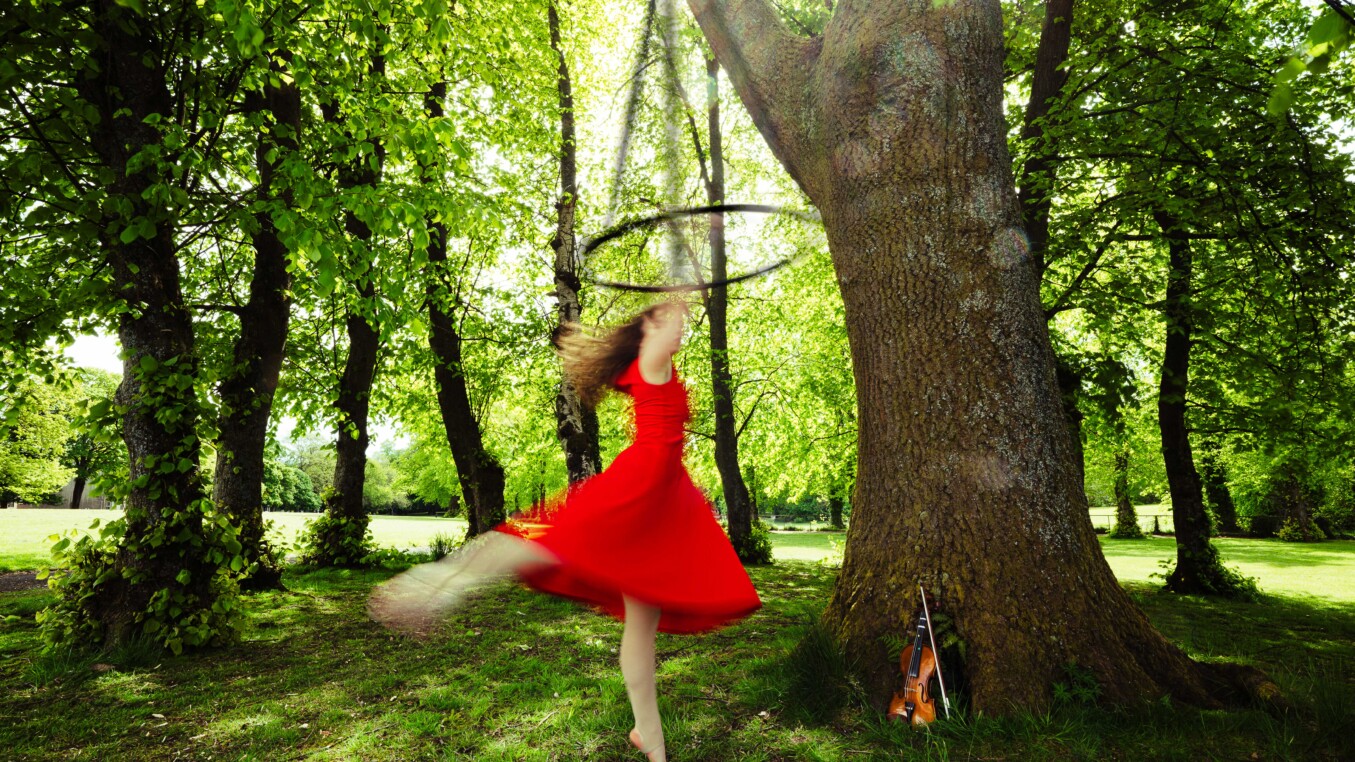A woman in a red dress is hanging from an aerial hoop, suspended from a tree.