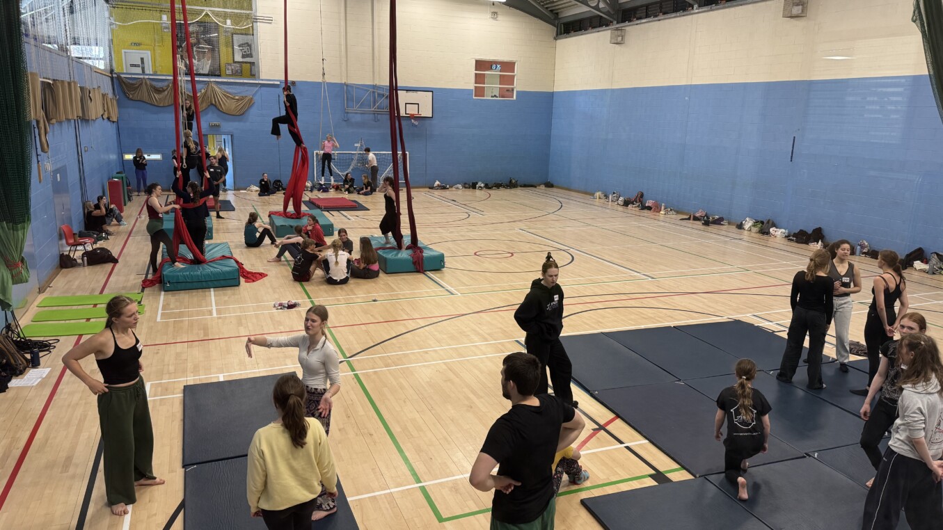 The image is of a sports hall, looking into it from an elevated position. There are lots of young people taking part in circus activities. In the foreground there are acrobatics, in the mid ground red silks, then at the back are people on trapeze
