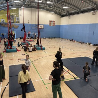 The image is of a sports hall, looking into it from an elevated position. There are lots of young people taking part in circus activities. In the foreground there are acrobatics, in the mid ground red silks, then at the back are people on trapeze