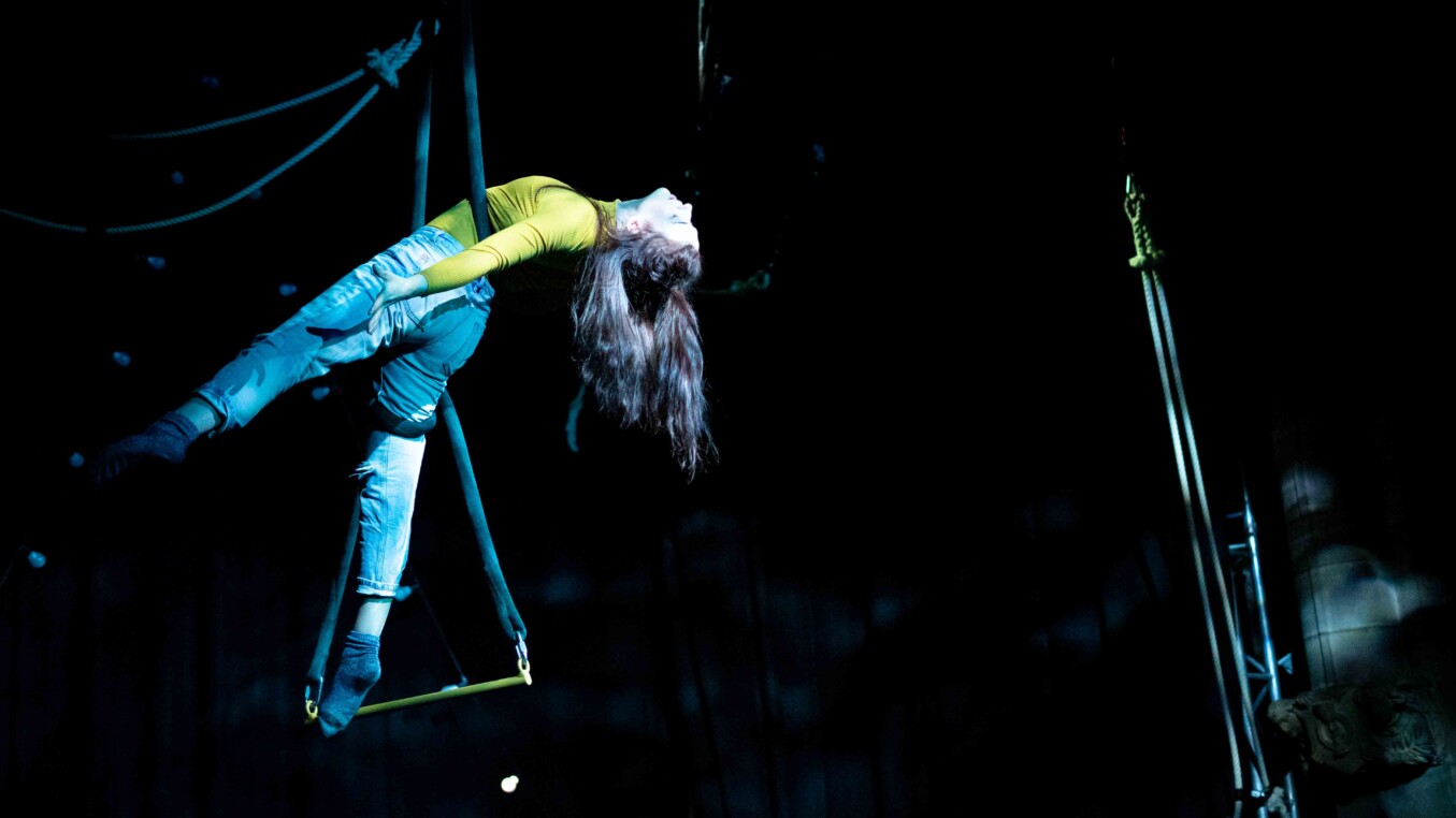A performer in a yellow top and jeans on an aerial trapeze mid-performance