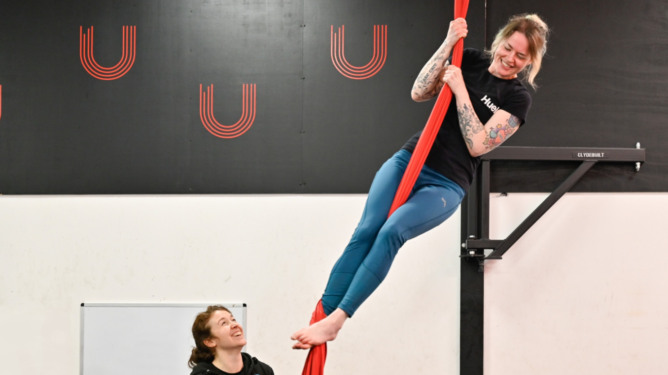 A woman in blue leggings standing upright on a red aerial silk while a tutor looks up at her. Her feet are at the tutor's head level.