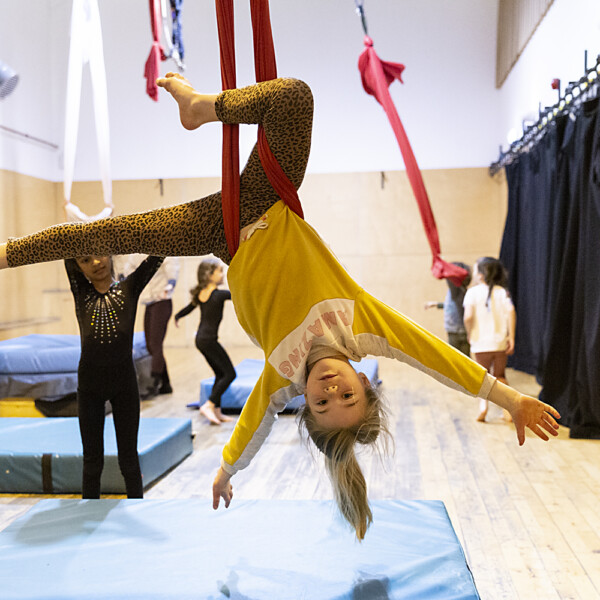 Girl in yellow jumper and leopard print leggings hangs upside down in an aerial silks hammock with her arms held wide
